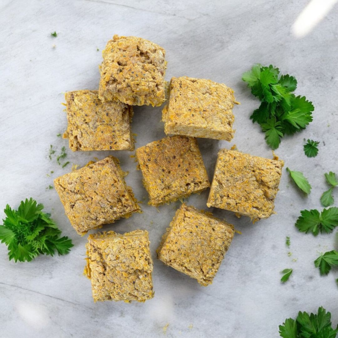 Square pieces of pork and pumpkin dog treats on a marble surface with fresh herbs